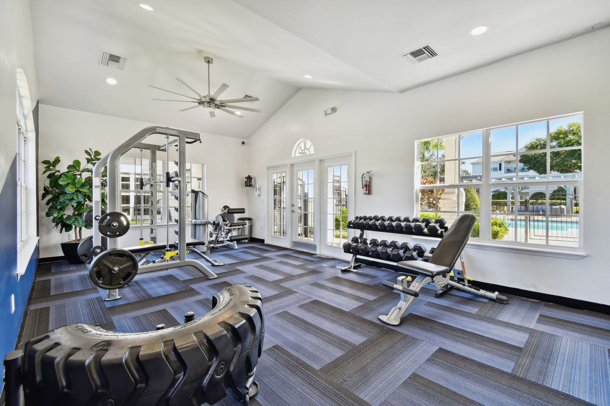 **Alt Text Description**: A well-equipped gym room with large windows letting in natural light and offering a view of an outdoor pool. Inside, the space features free weights on a rack with an adjustable weight bench positioned in front, a multi-station cable machine with weight plates, and a large tractor tire for functional fitness training. The flooring consists of blue and black patterned carpet tiles, and a ceiling fan is mounted above to keep the area cool. The bright white walls and clean design contribute to a fresh and modern atmosphere.