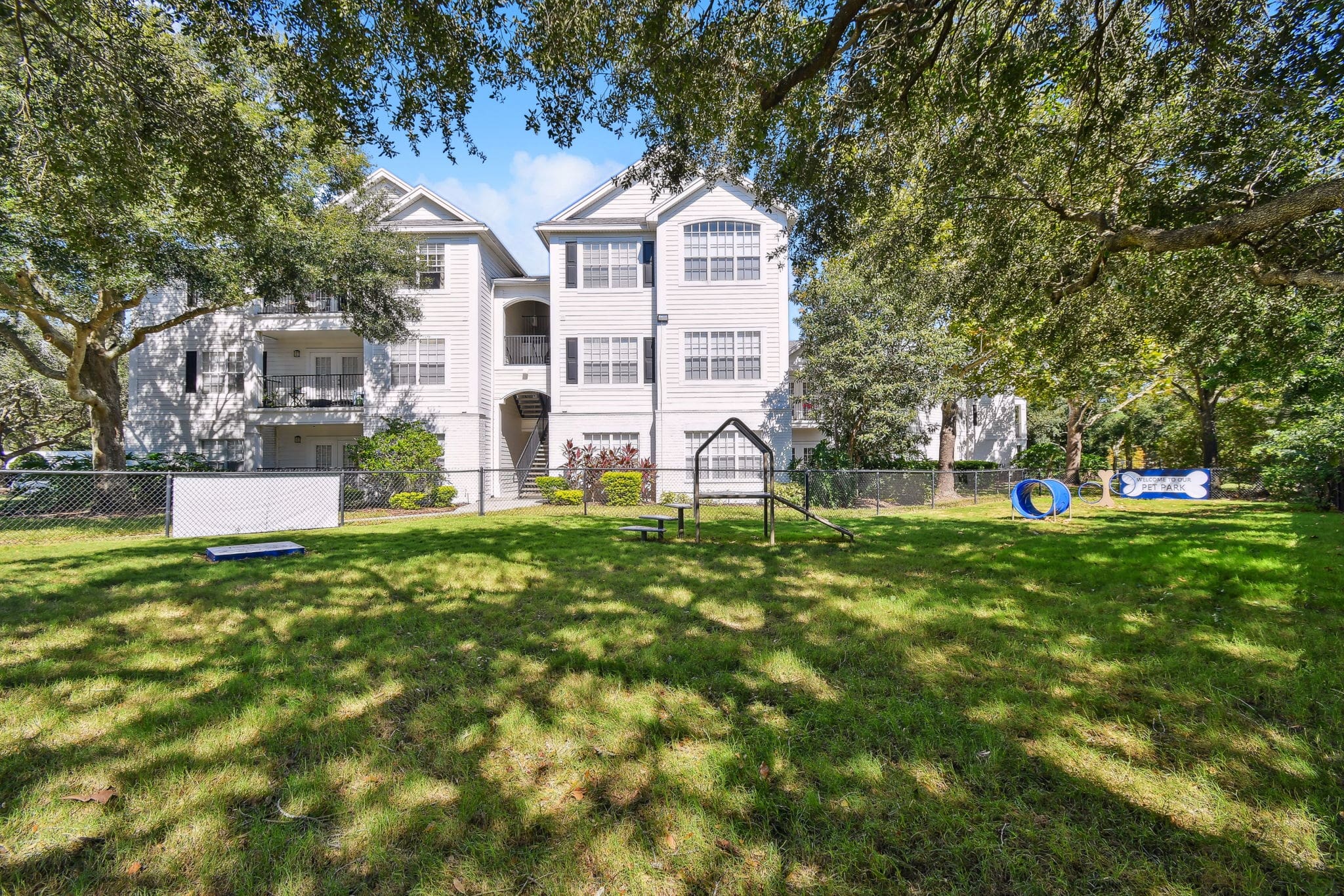 **Alt Text Description**: A spacious, grassy pet park surrounded by a chain-link fence, located behind a white, three-story residential building. The park includes agility equipment such as a blue tunnel, a ramp, a balance beam, and a small platform. Tall trees provide ample shade over the area, and a sign in the shape of a bone reads “Pet Park.” The well-maintained lawn is dappled with sunlight filtering through the tree canopy.