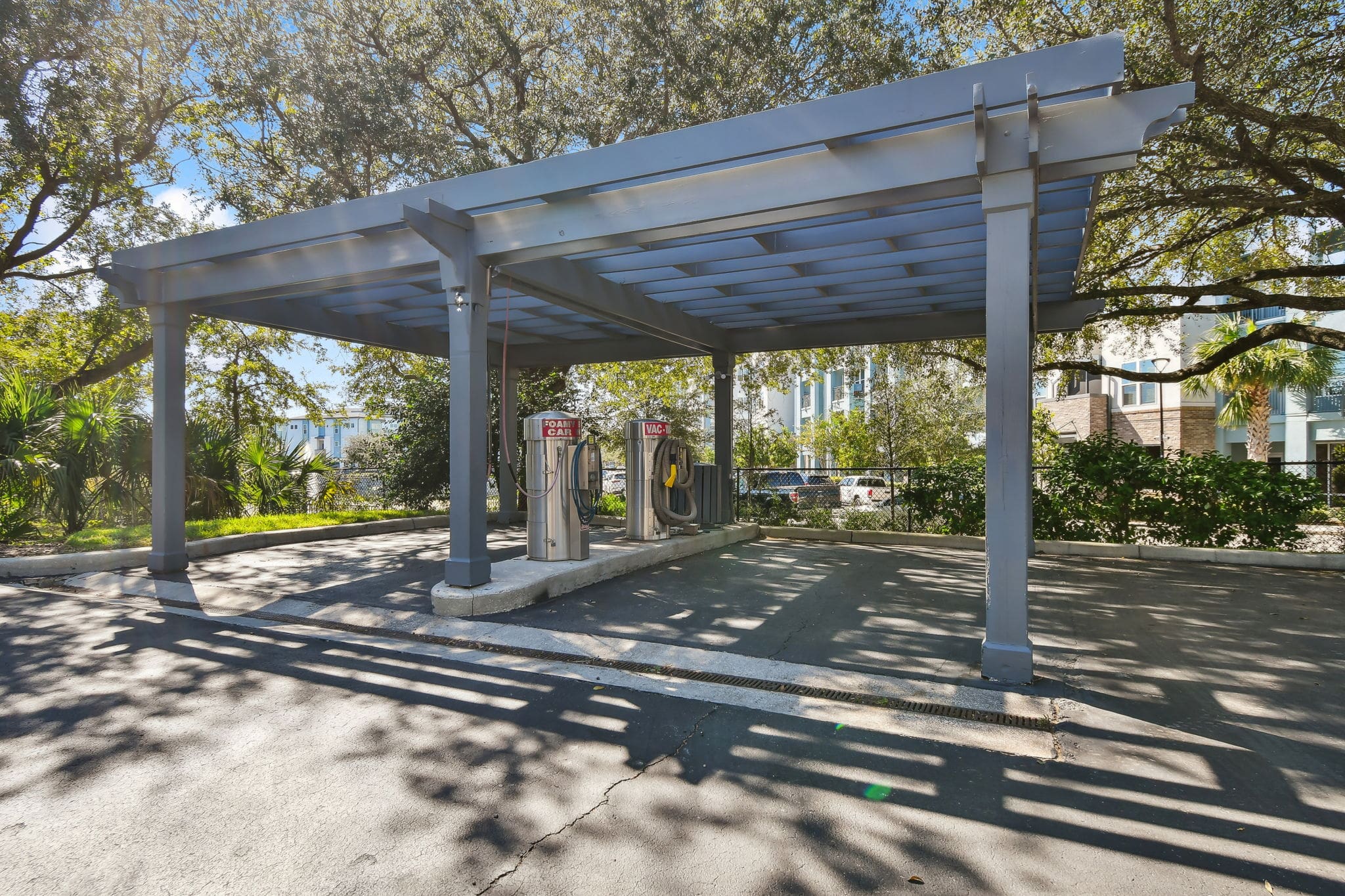 **Alt Text Description**: A shaded car care station with a pergola-style roof supported by large gray wooden beams. Under the structure are two stainless steel machines labeled “Foam Carpet Cleaner” and “Vacuum,” each equipped with coiled hoses and nozzles. The station is built on a concrete island in a black asphalt parking area, surrounded by lush greenery, palm trees, and a black chain-link fence. Residential buildings are partially visible through the foliage in the background.