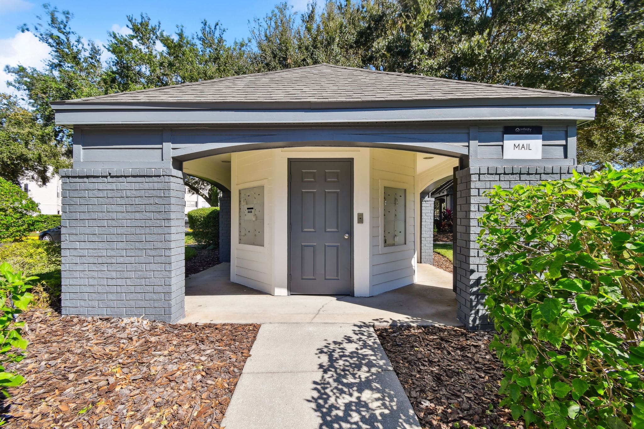 **Alt Text Description**: A small, standalone mail kiosk structure with a peaked roof, gray brick pillars, and white wooden paneling. A central door is flanked by multiple mailbox compartments on both visible side walls. A sign reading "MAIL" is mounted above the entrance. The structure is set within a landscaped area with mulch, green shrubs, and trees under a clear blue sky.